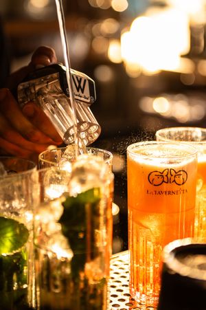 Bartender pouring a cocktail into glasses at La Tavernetta bar, with warm golden bokeh lights in the background.