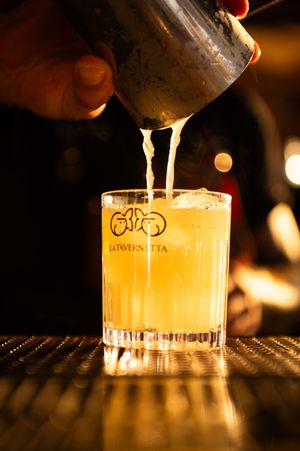 Bartender pouring a golden cocktail into a branded La Tavernetta rocks glass on a illuminated bar mat.