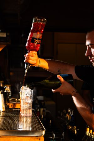 Bartender pouring Aperol into a tall glass with ice at a dimly lit bar.