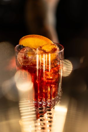 A vibrant red Negroni cocktail garnished with an orange slice, served over ice in a crystal glass with warm bokeh lighting.