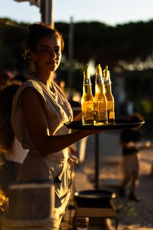 A waitress holds a tray of Corona beer bottles outdoors at golden hour, backlit by warm sunset light.