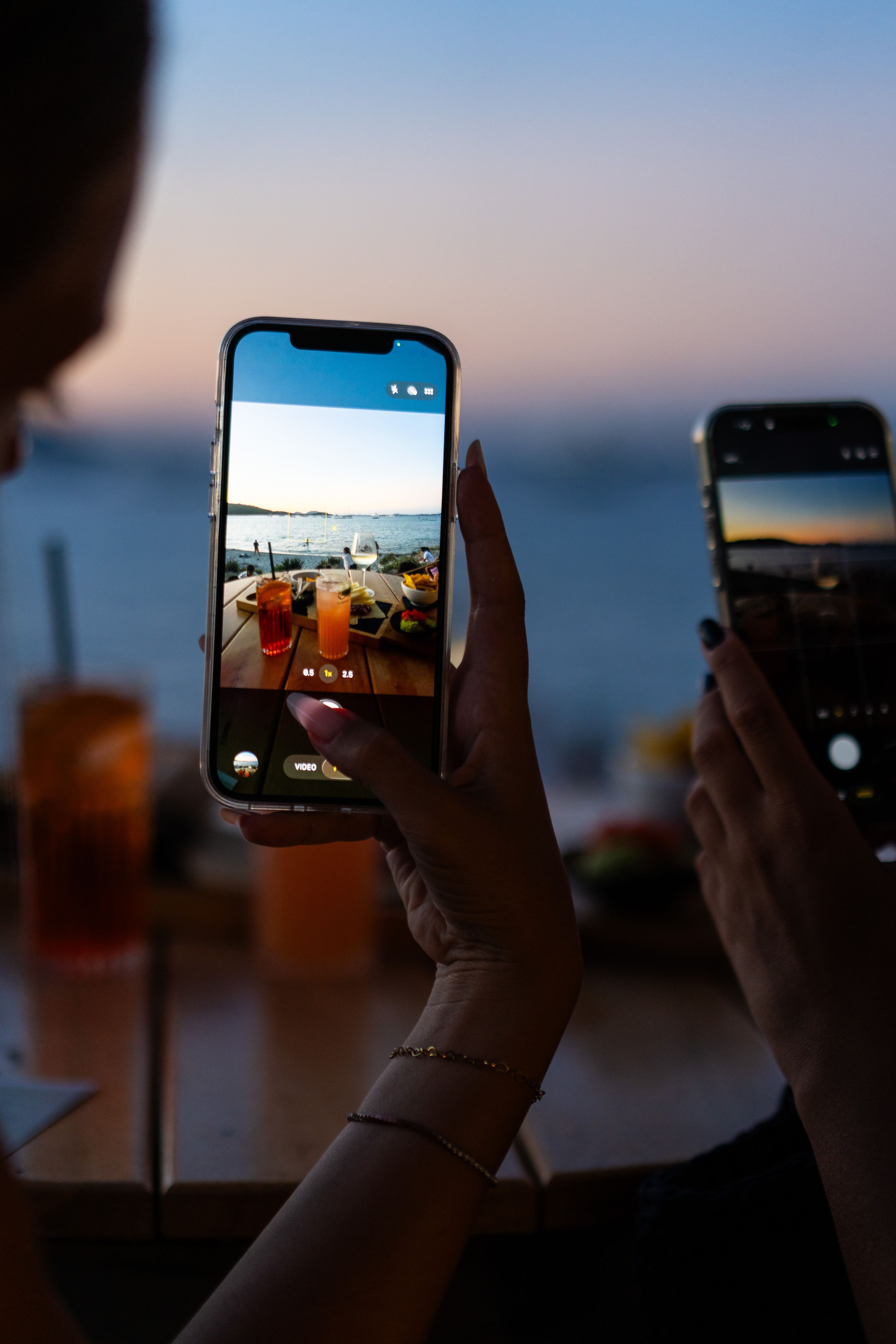 Two women photograph cocktails on smartphones at a seaside bar during a colorful sunset evening.