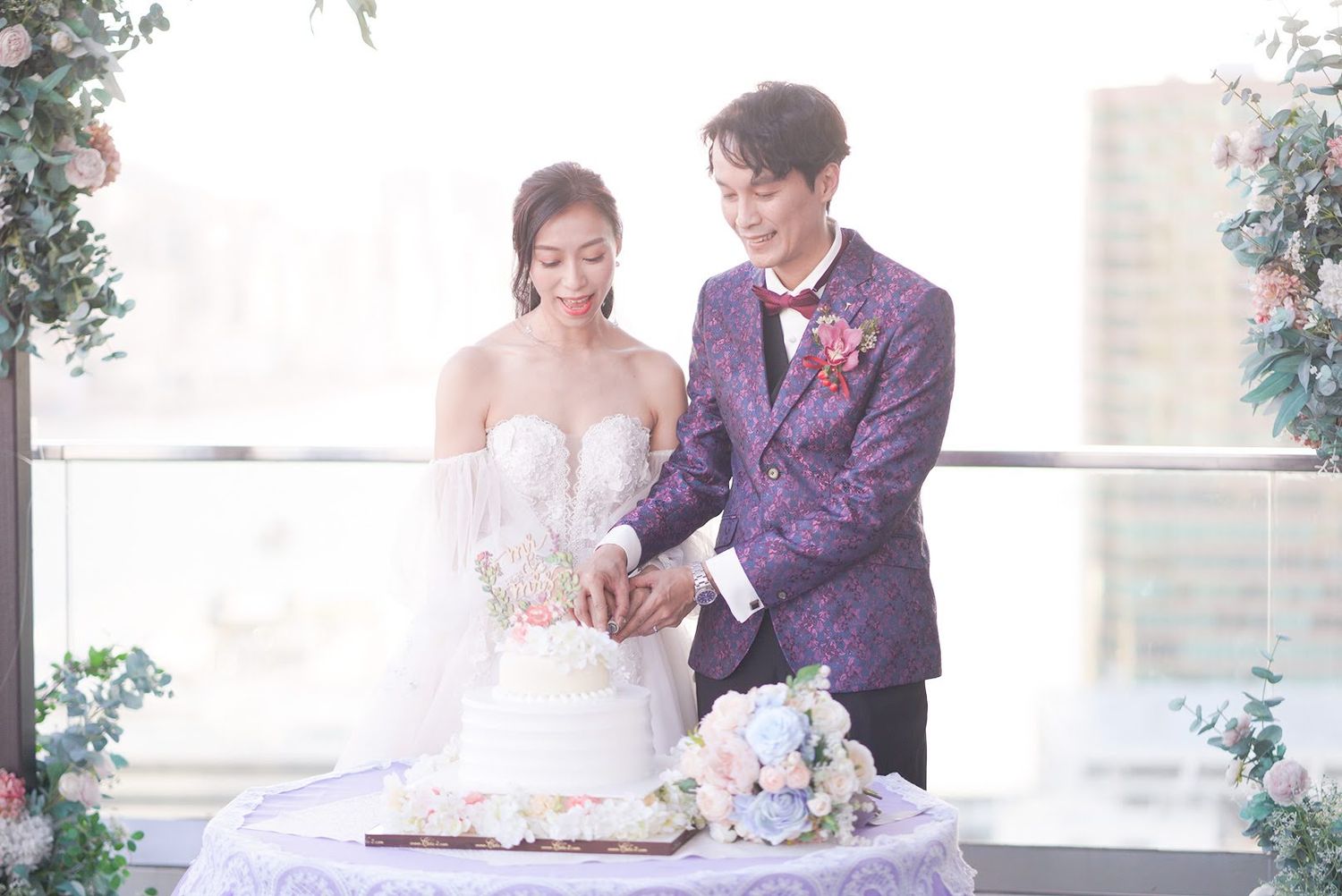 A couple in wedding attire stands together at an elegant reception cutting their white tiered wedding cake.