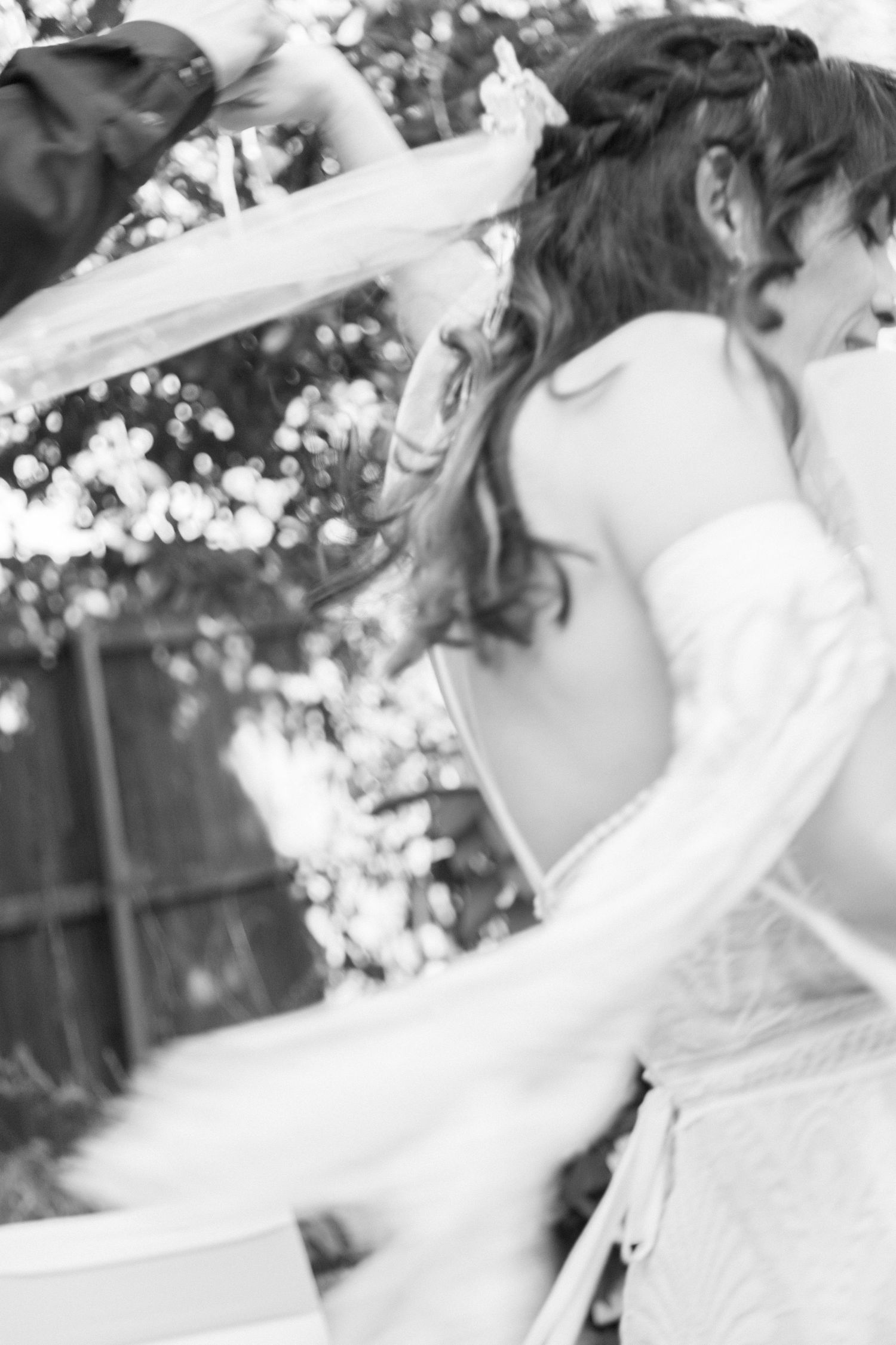 Black and white photo of a bride in a white gown dancing joyfully outdoors, veil flowing, with bokeh lights in background.