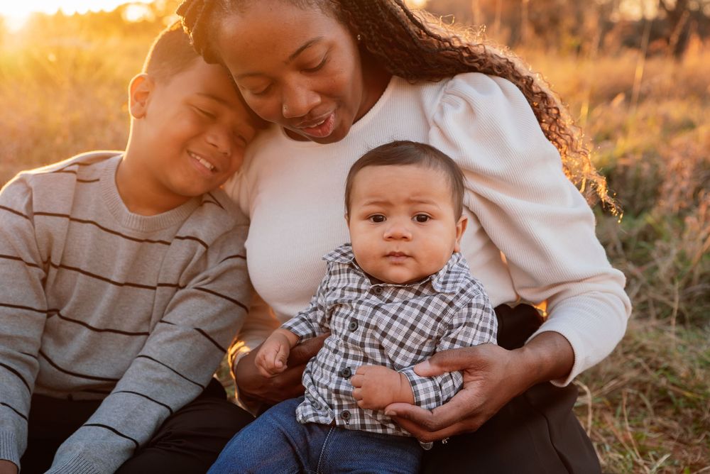 A mother and two children sit together in a golden sunset field, smiling warmly in a heartfelt family portrait.