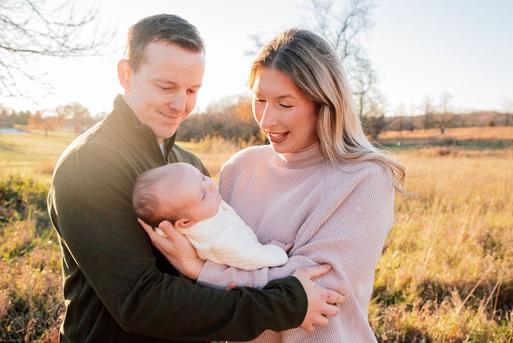 A couple smiles at their newborn baby outdoors in a golden sunlit field during a family portrait session.