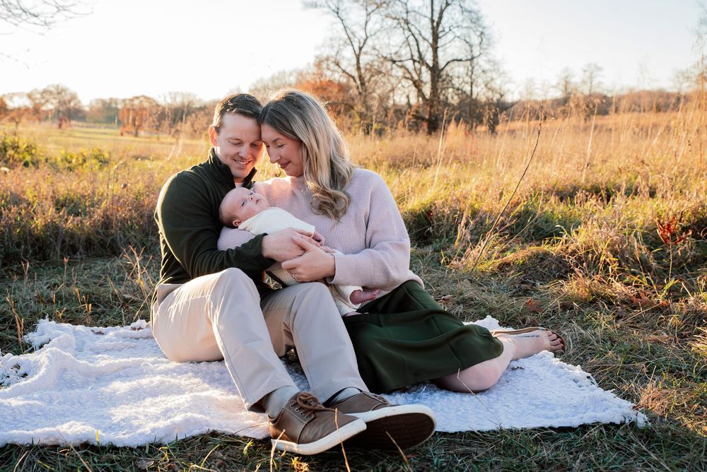 Couple sitting on blanket in autumn field holding newborn baby during golden hour family photo session.