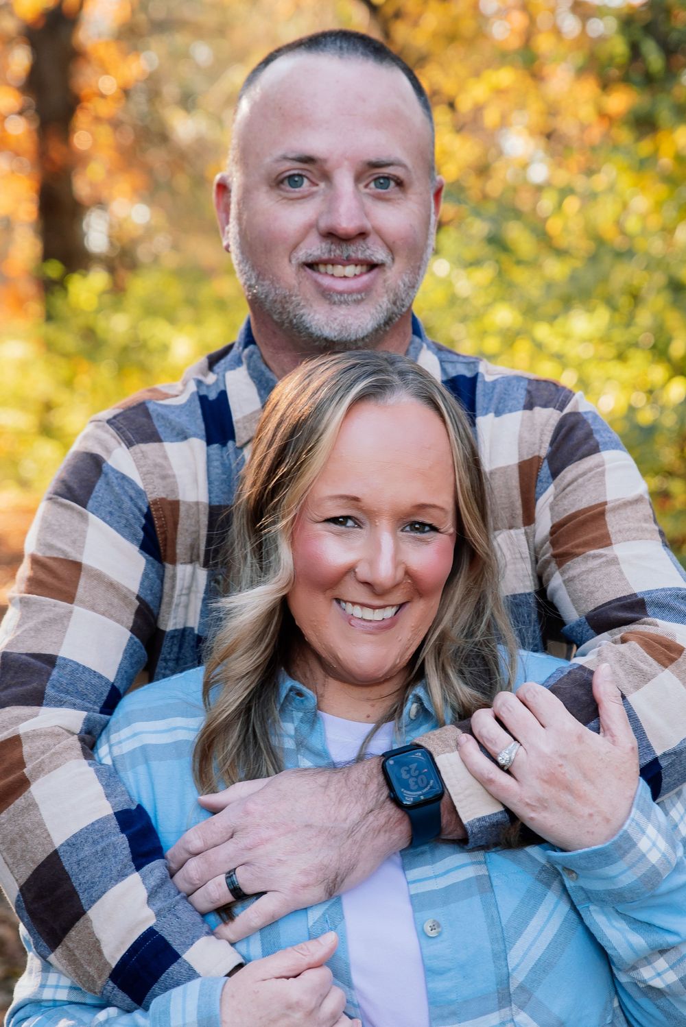 Smiling couple in matching plaid shirts embracing outdoors with colorful autumn foliage in the background.