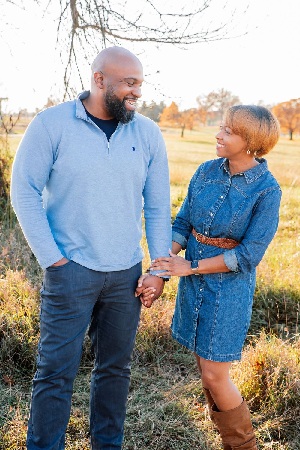 A smiling couple holds hands in a sunlit autumn field, man in blue pullover and woman in denim dress with brown boots.