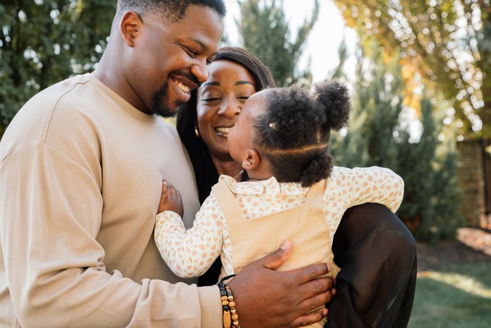 Happy Black family with parents smiling and hugging their young daughter outdoors in a sunny garden setting.