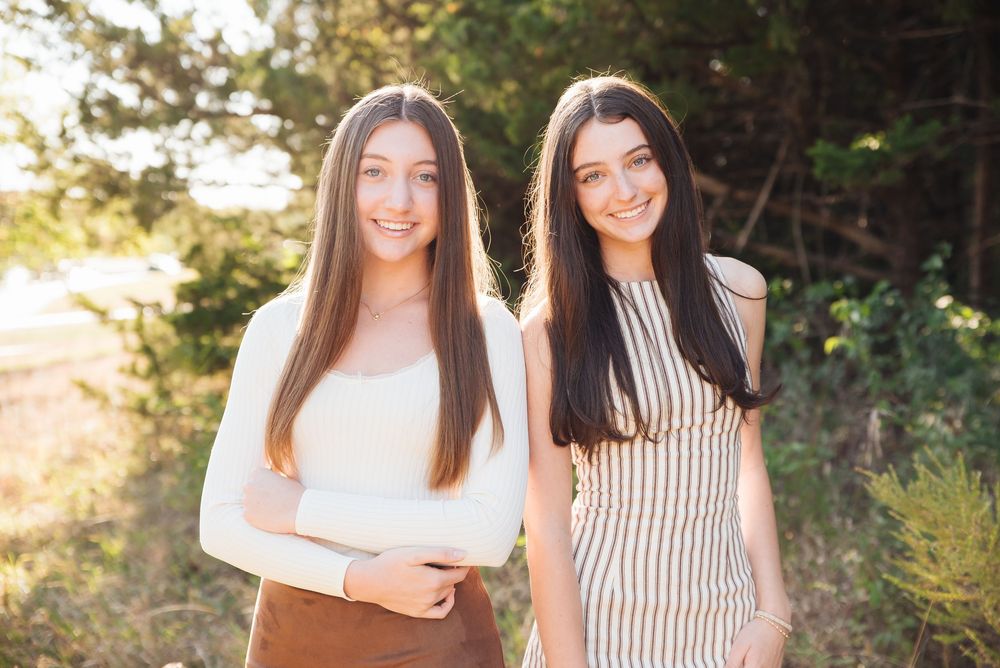 Two smiling young women with long dark hair pose outdoors in warm golden sunlight near green trees.