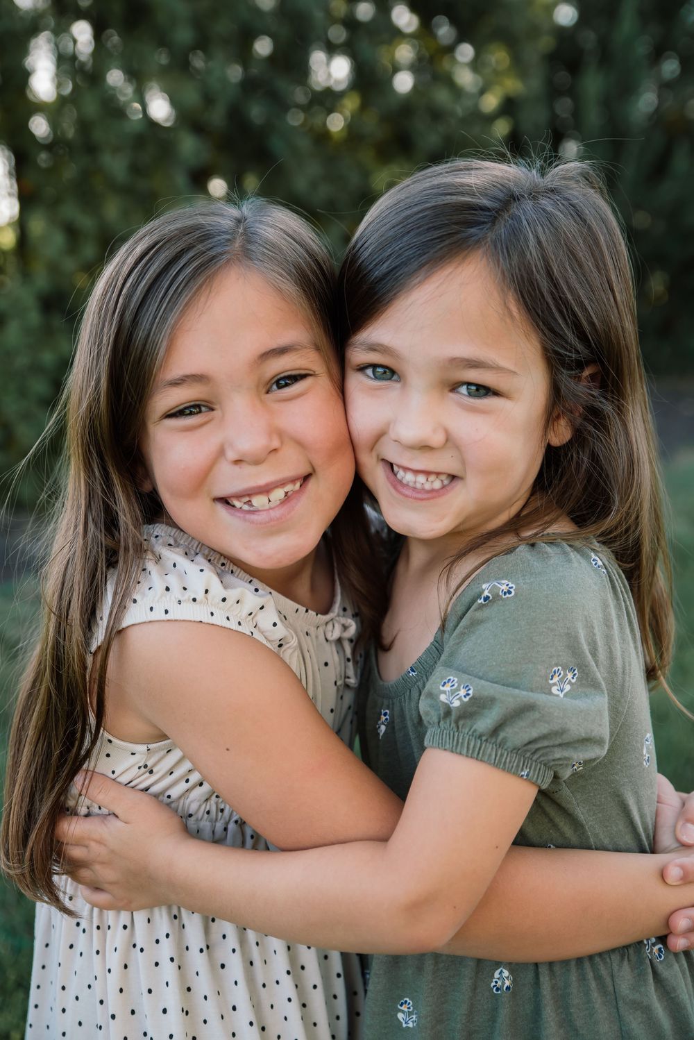 Two young girls with brown hair smiling and hugging outdoors in a green park setting.