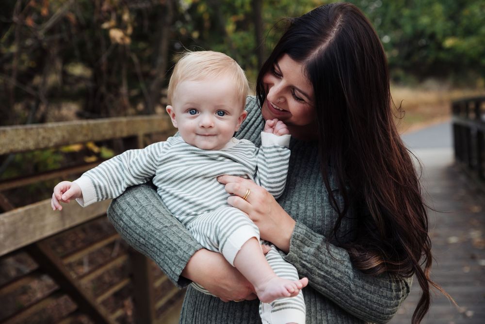 A smiling mother with long dark hair holds her happy baby on a wooden bridge surrounded by autumn foliage.