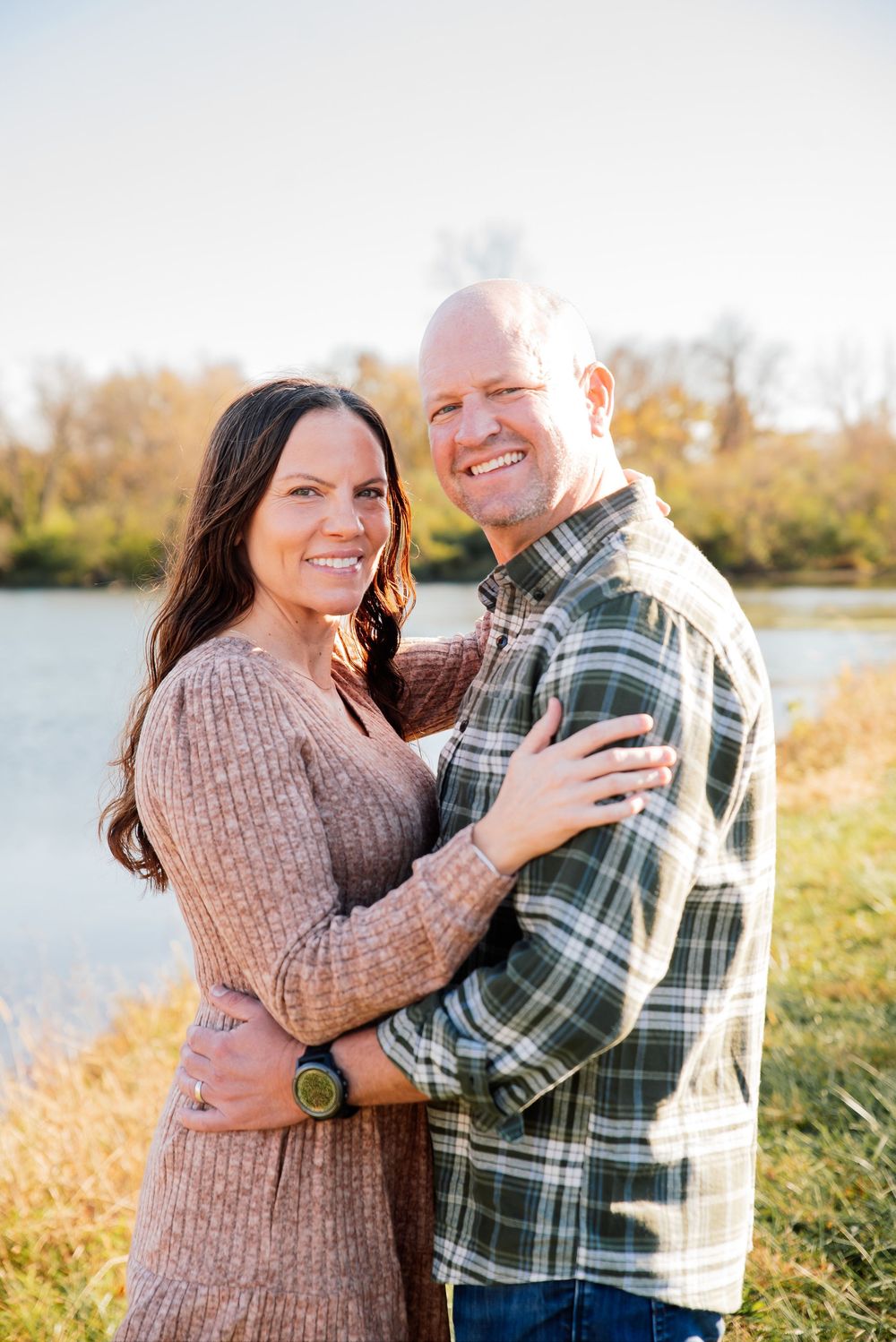 A smiling couple embraces outdoors near a lake in autumn, wearing casual fall attire in warm golden light.