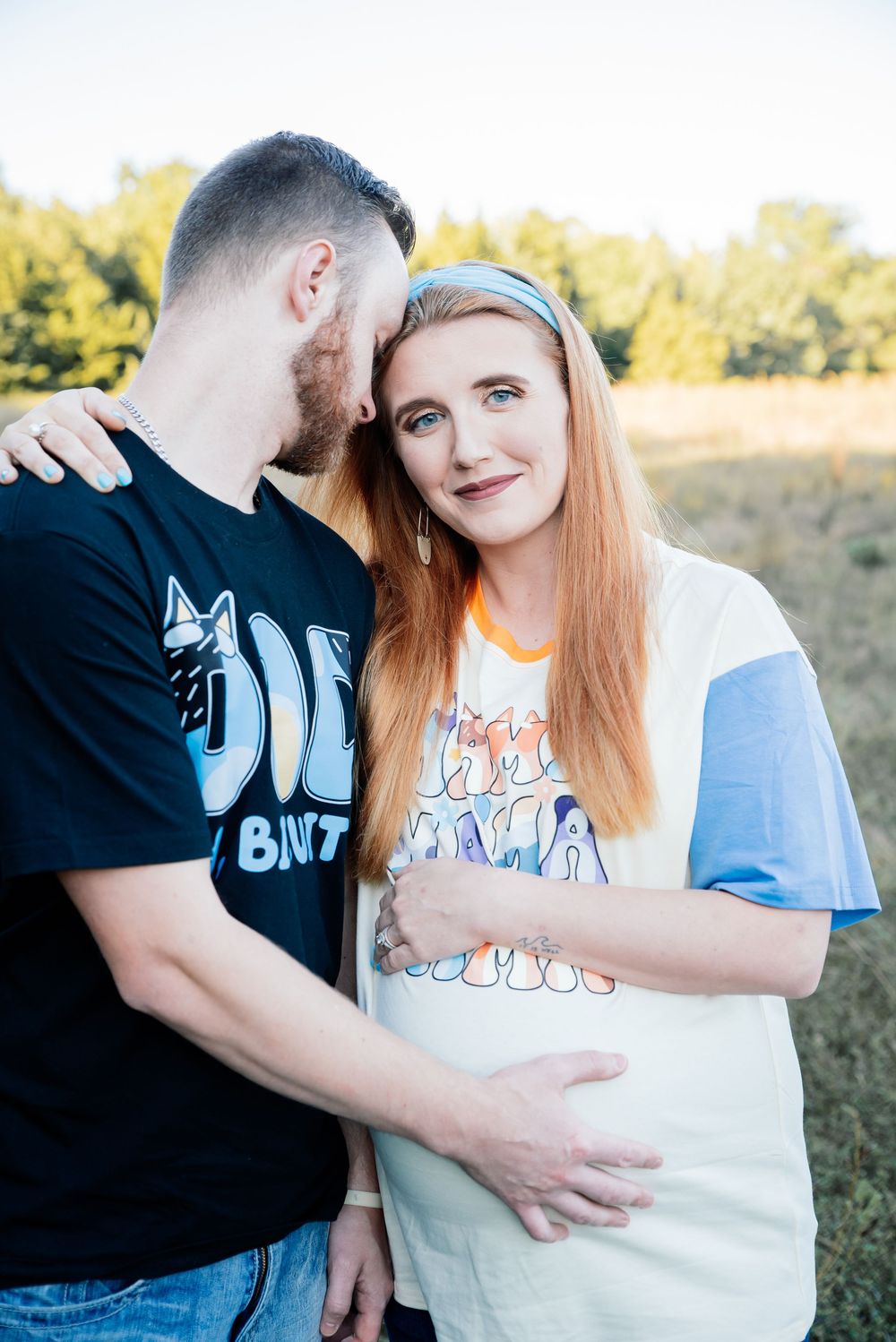 A pregnant woman in a colorful t-shirt stands with her partner outdoors, both smiling in a sunny field.
