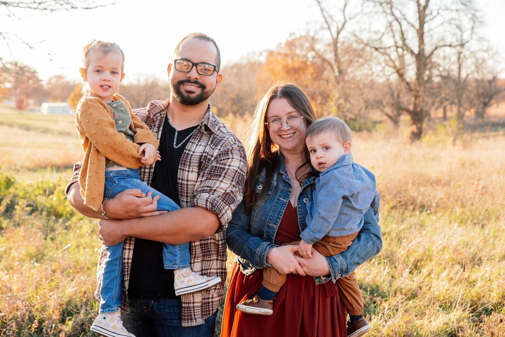 A happy family of four poses in a golden autumn field, with parents each holding a young toddler outdoors.