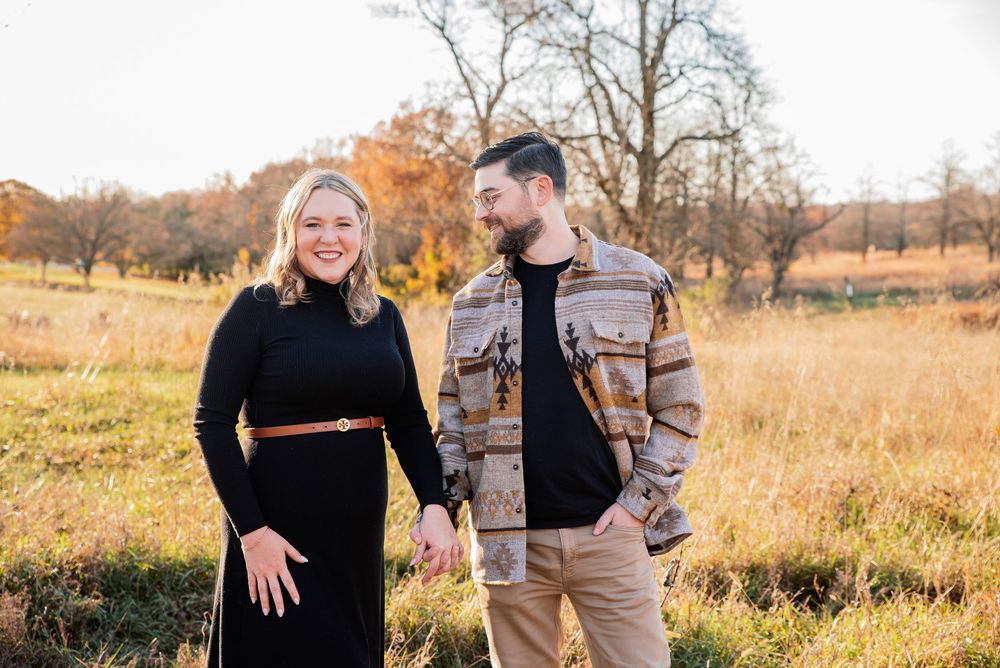 Couple holding hands smiling at each other in a golden autumn field with colorful fall foliage trees.