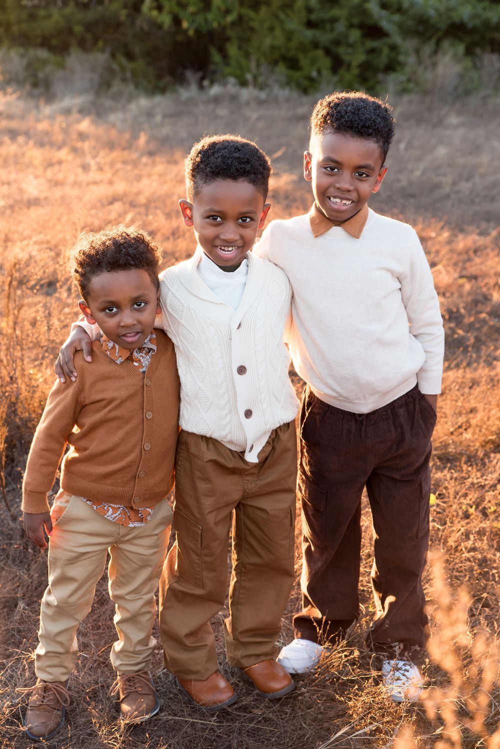 Three young boys smiling and posing together outdoors in a golden field, dressed in coordinated earth-tone outfits.