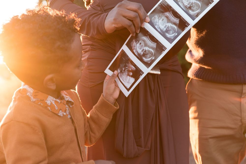 Child examining ultrasound photos held by pregnant mother in warm golden sunset light outdoors.