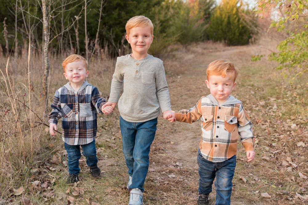 Three red-haired boys holding hands walk along a dirt path surrounded by autumn foliage in a family portrait.