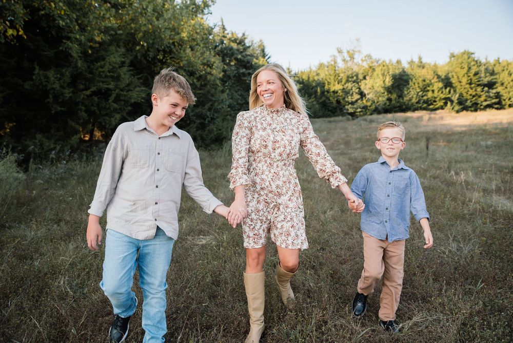 A woman in a floral dress holds hands with two boys walking through a sunny green field.