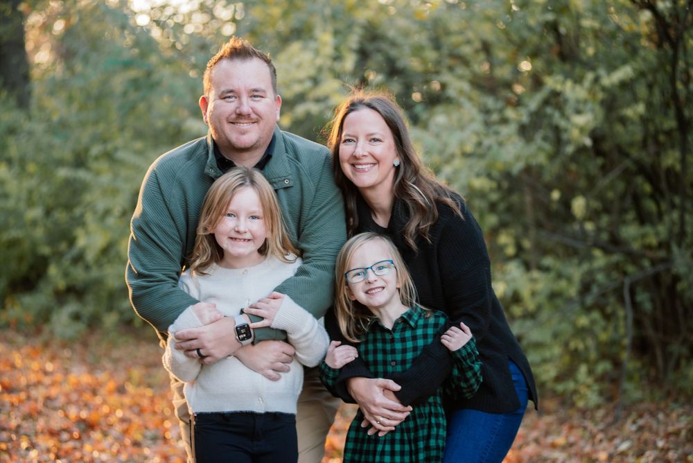 Family of four smiling outdoors in autumn, parents with two young girls wearing glasses, surrounded by fall foliage.