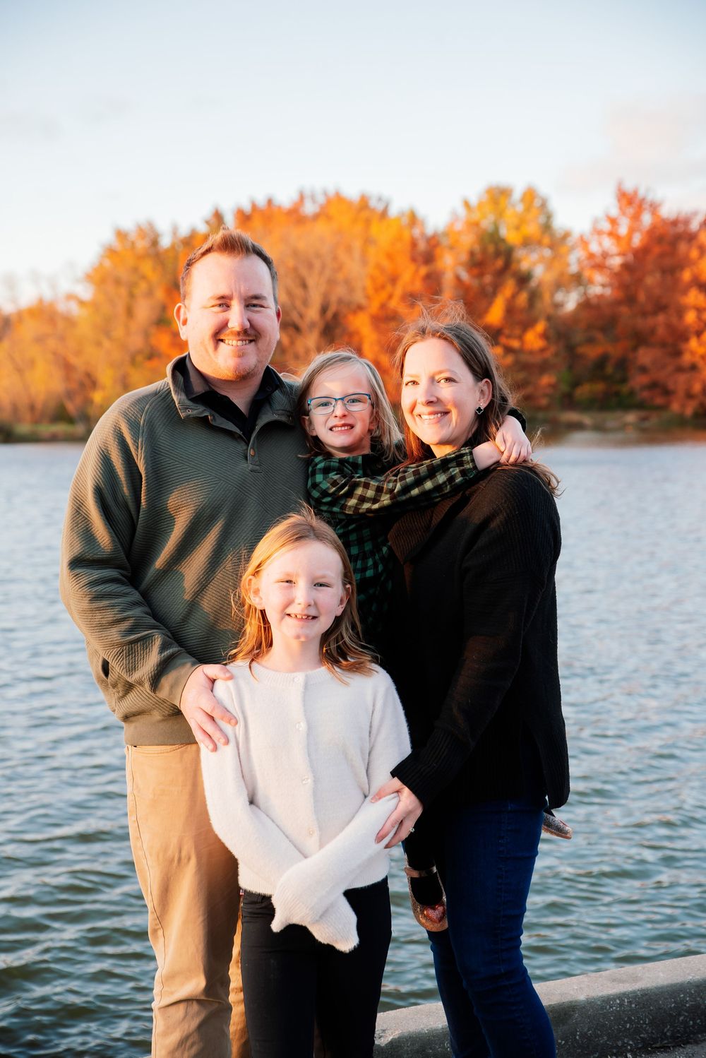 Family of four smiling by a lake with vibrant autumn foliage in the background on a sunny fall day.