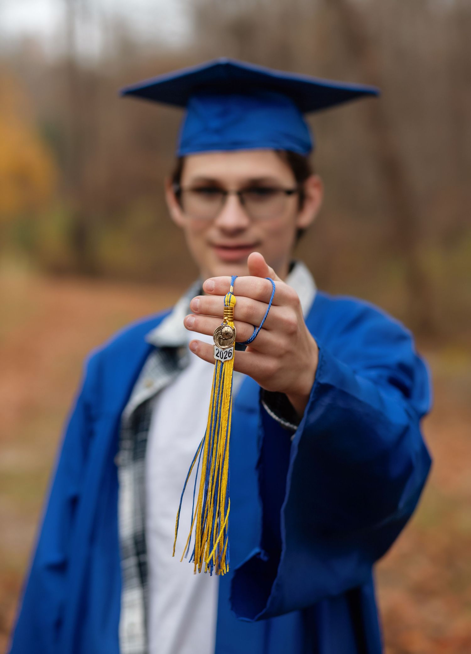Young graduate in blue cap and gown holds tassel toward camera outdoors in autumn setting.