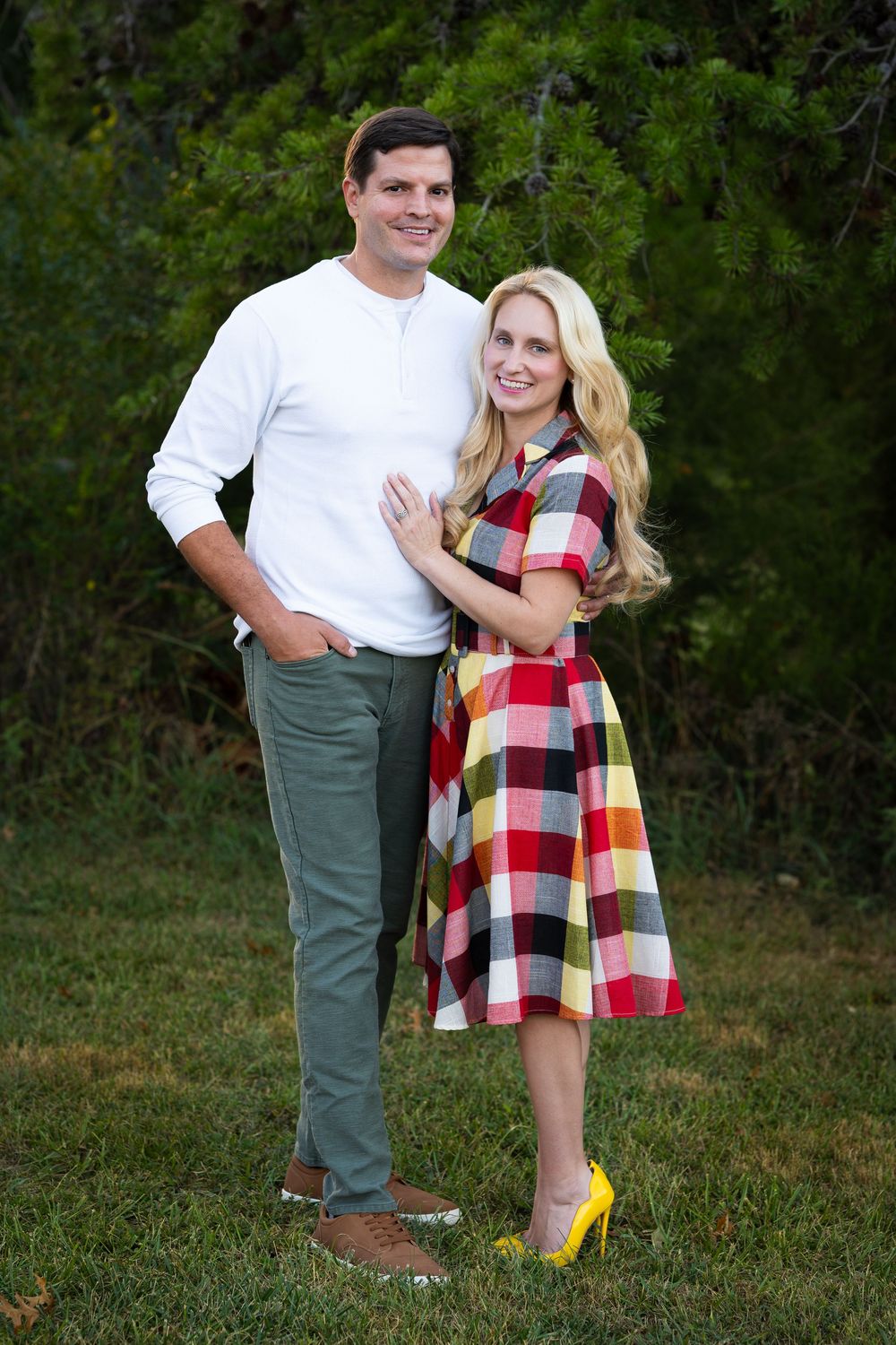 A smiling couple poses outdoors, woman in plaid dress and man in white shirt.