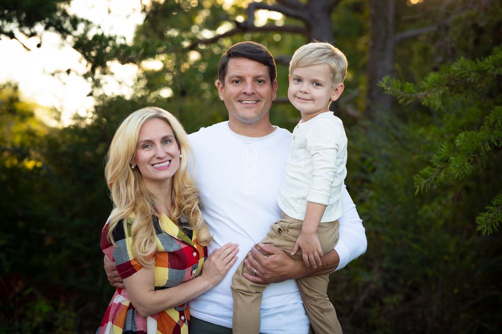A smiling family of three poses outdoors at golden hour among green trees.