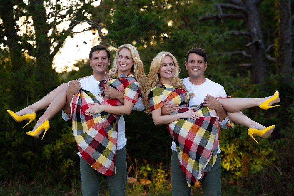 Two men carrying two blonde women wearing matching plaid dresses and yellow heels outdoors.