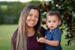 Smiling mother posing with young toddler son holding a leaf near magnolia tree outdoors.