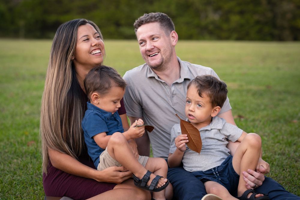 Joyful family of four sitting on grass laughing together in sunny outdoor park setting.