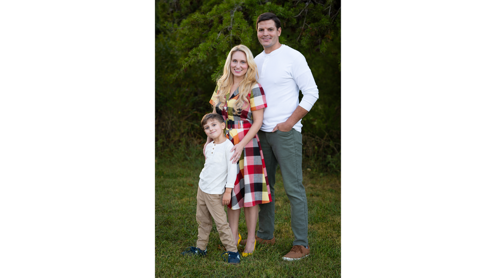 Family of three posing together outdoors, mother in plaid dress with husband and young son.