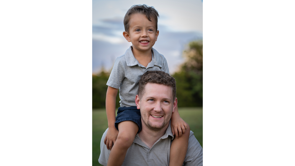 Smiling father with young son sitting on his shoulders in outdoor green field setting.