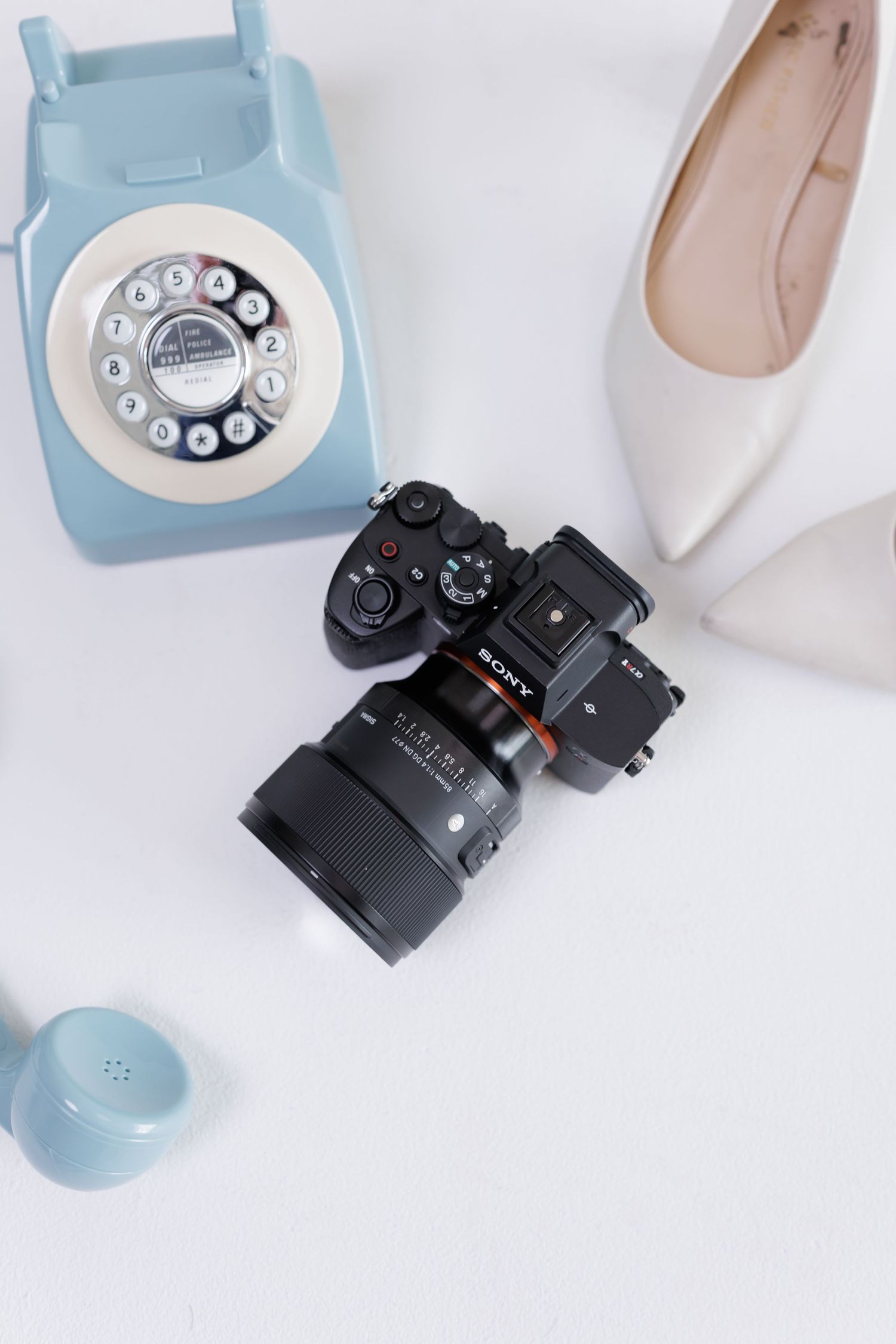 Flat lay of Sony mirrorless camera beside vintage blue rotary phone and beige heels on white surface.