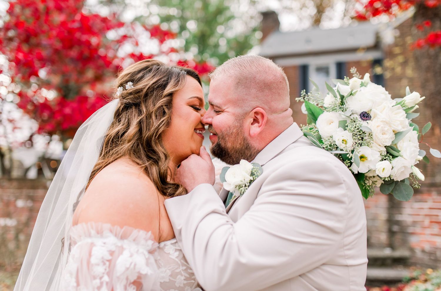 Bride and groom share a romantic moment, bride holding white floral bouquet, red foliage backdrop.