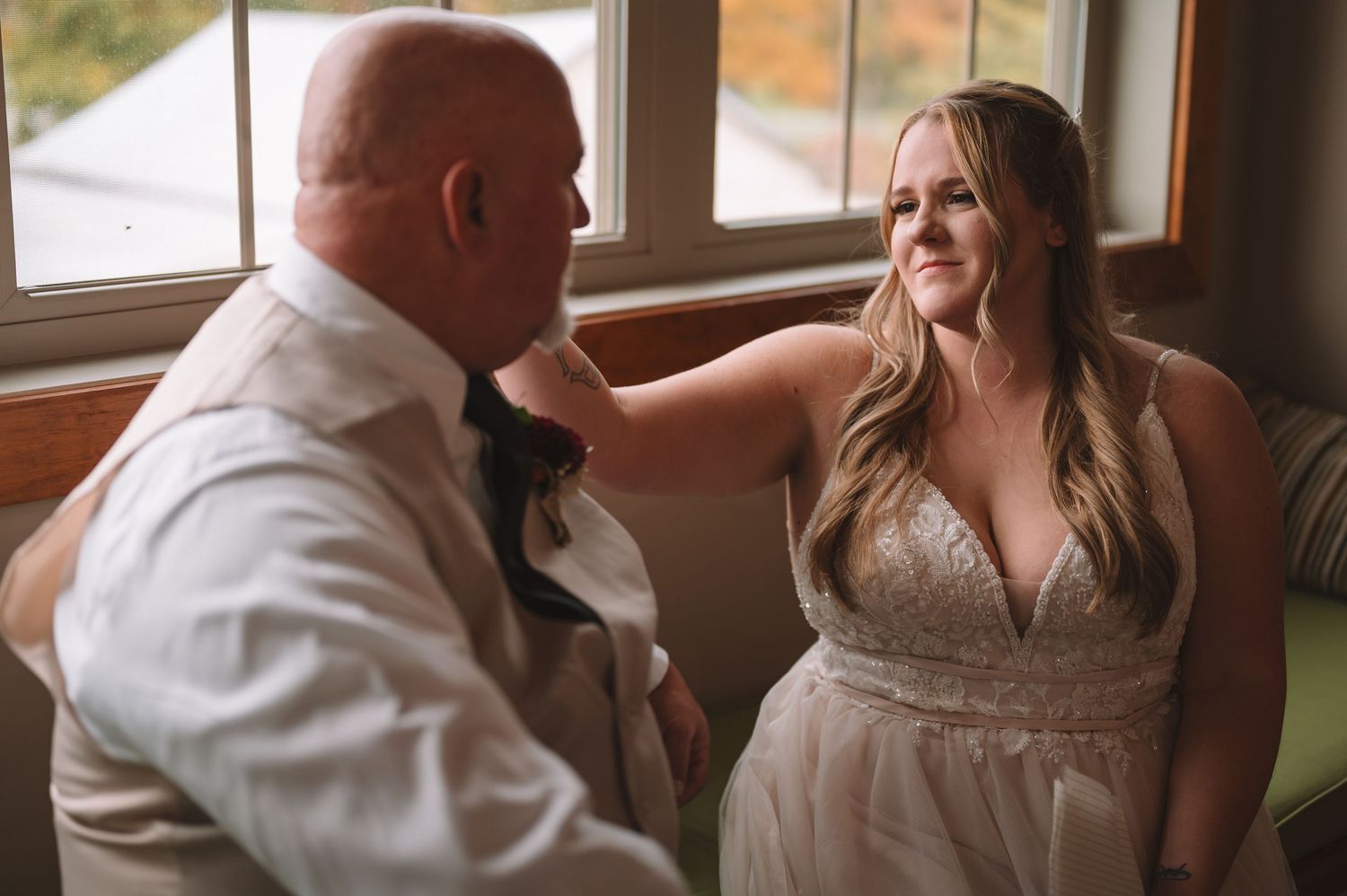Bride in white lace gown smiles at father touching his face near window on wedding day.