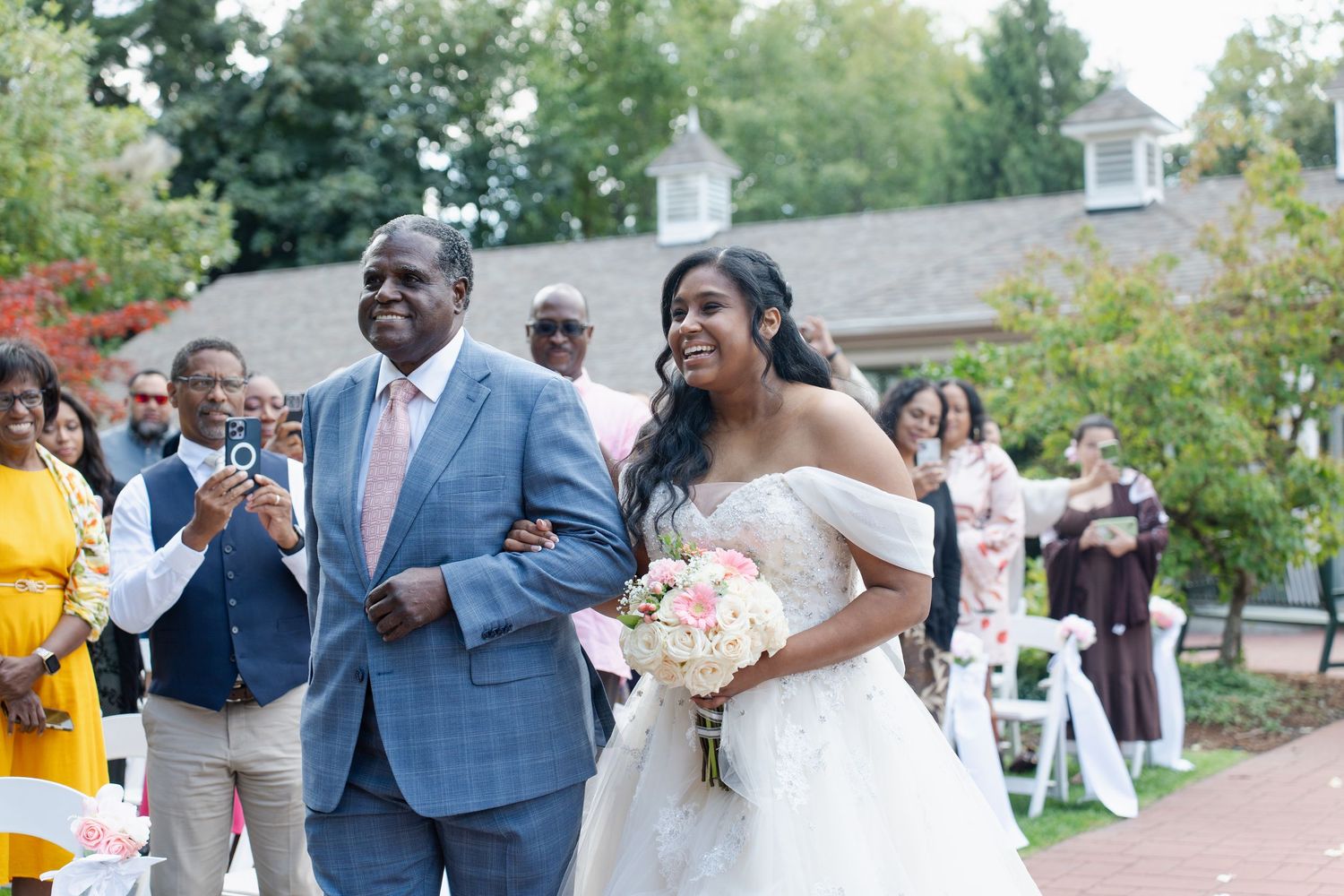Bride in white off-shoulder gown walks down aisle with escort at outdoor wedding ceremony.