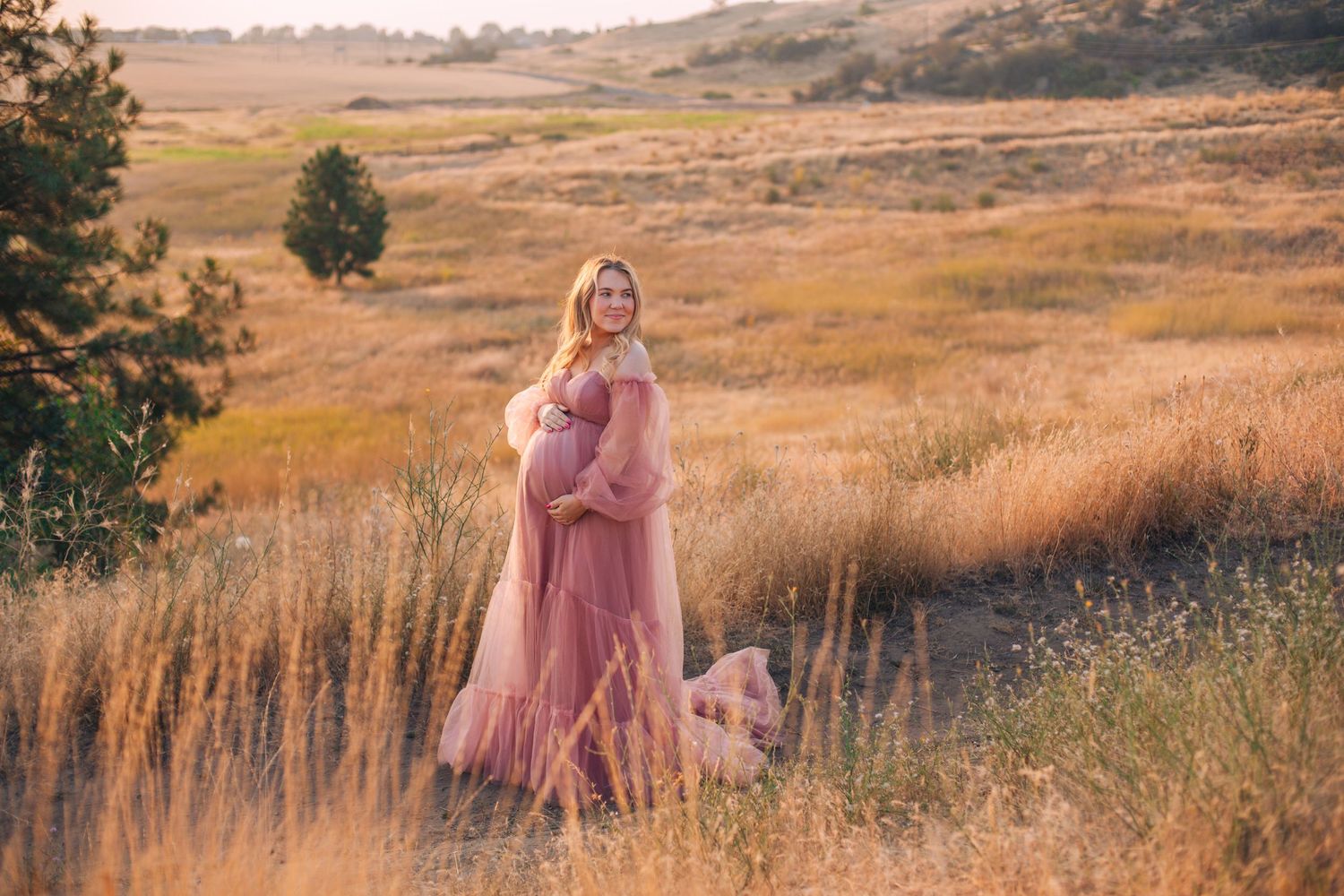 Pregnant woman in pink flowing maternity gown posing in golden field at sunset during outdoor maternity photo session.
