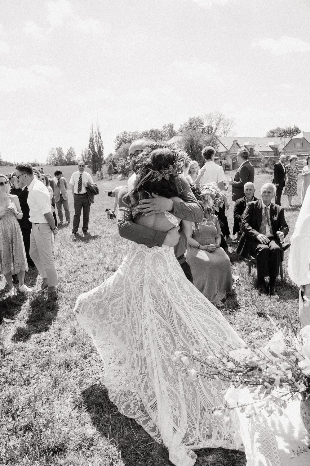 Black and white photo of a bride in lace gown embracing someone at an outdoor wedding ceremony with guests seated nearby.