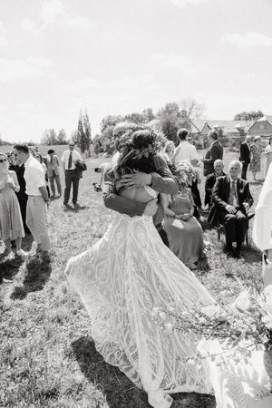 Black and white photo of a bride in lace gown embracing someone at an outdoor wedding ceremony with guests seated nearby.