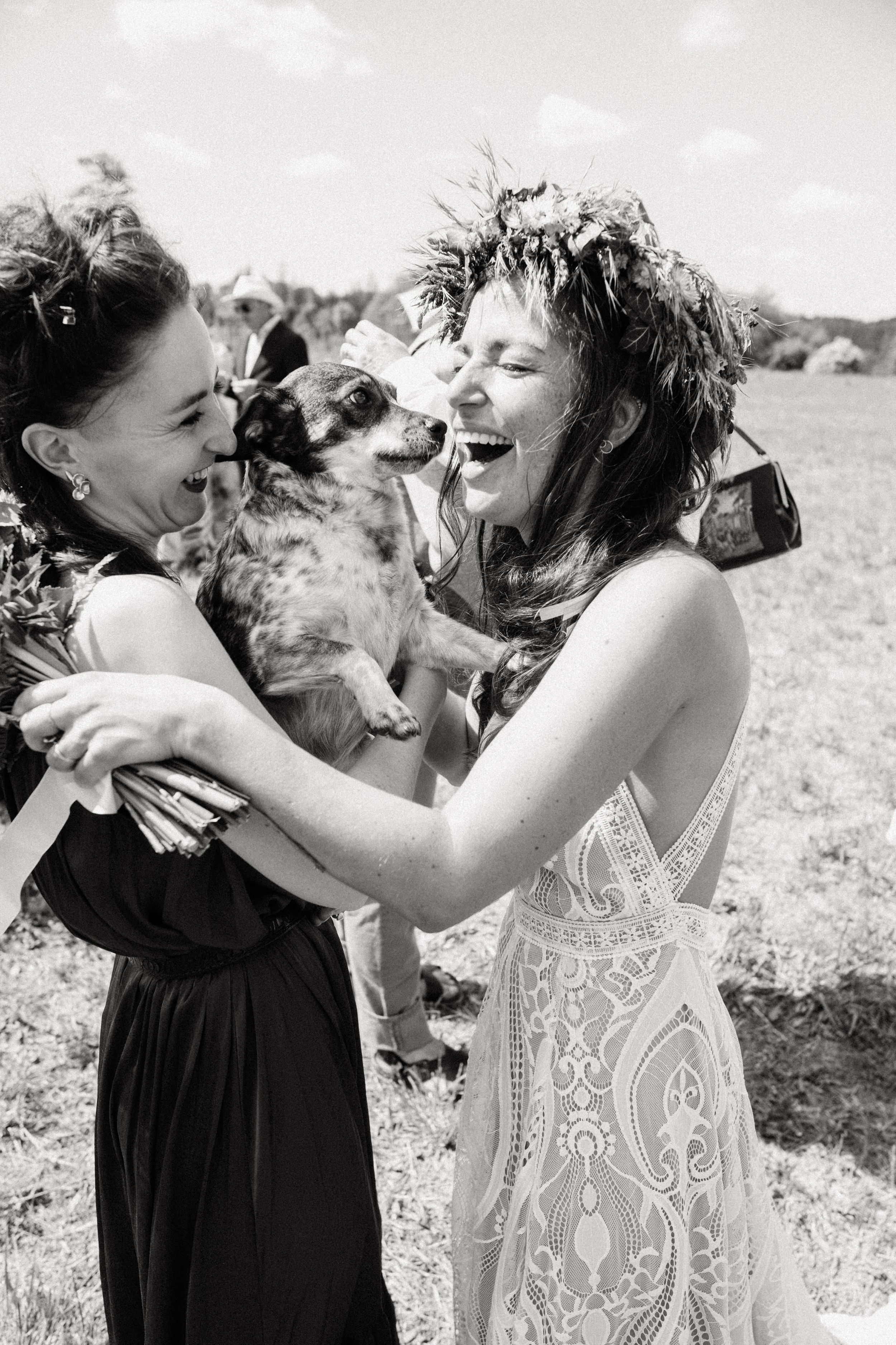 Black and white photo of two women laughing with a dog at an outdoor wedding celebration.