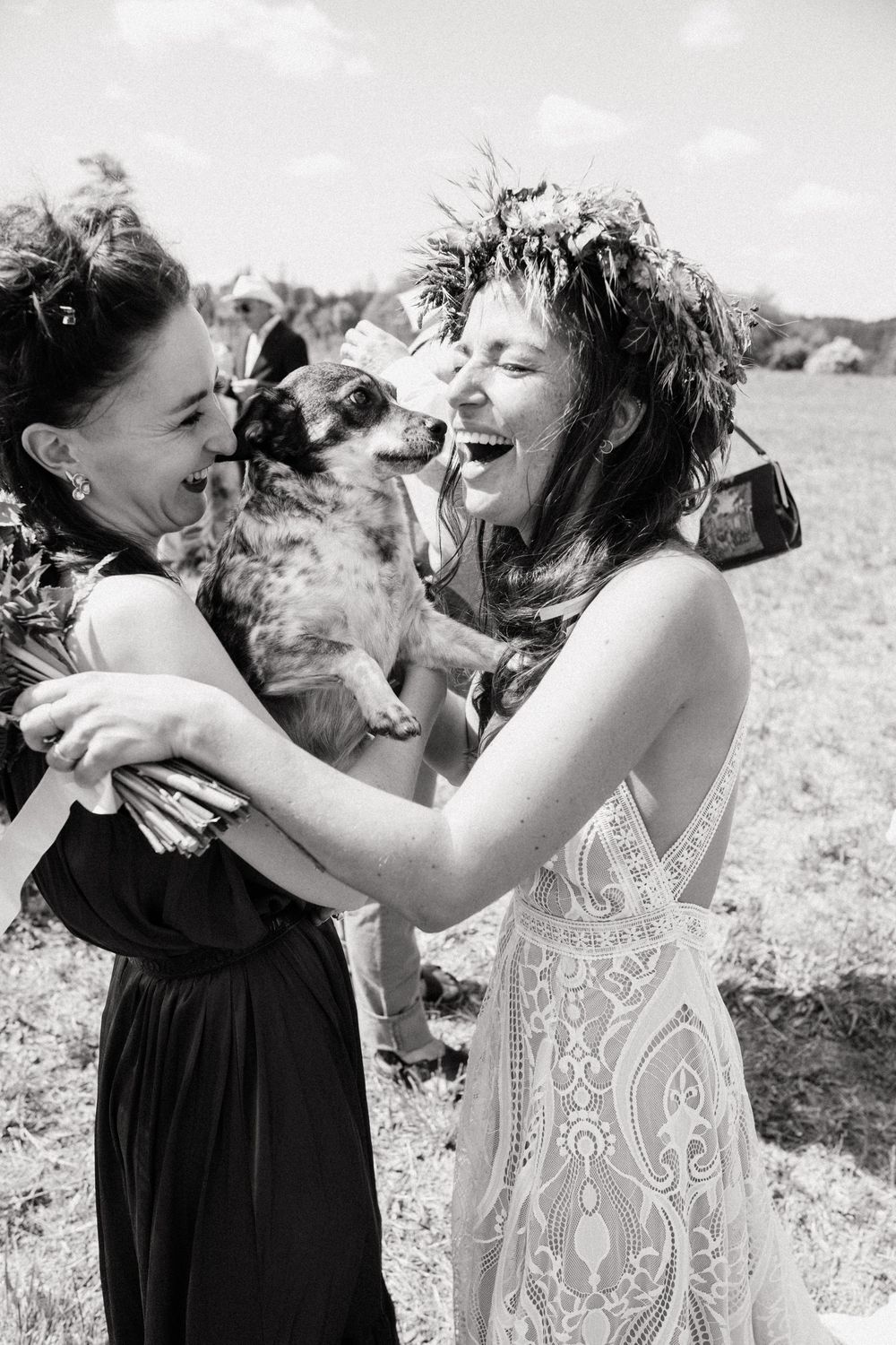 Black and white photo of two women laughing with a dog at an outdoor wedding celebration.