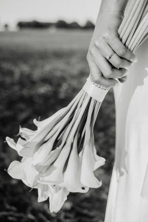 Black and white photo of a bride holding calla lily bouquet with ring and bracelet visible, field in background.
