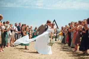 Bride and groom share a romantic dip kiss at outdoor wedding ceremony with guests applauding under blue sky.