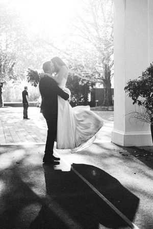 Black and white photo of a bride and groom sharing a kiss outdoors near a column on their wedding day.
