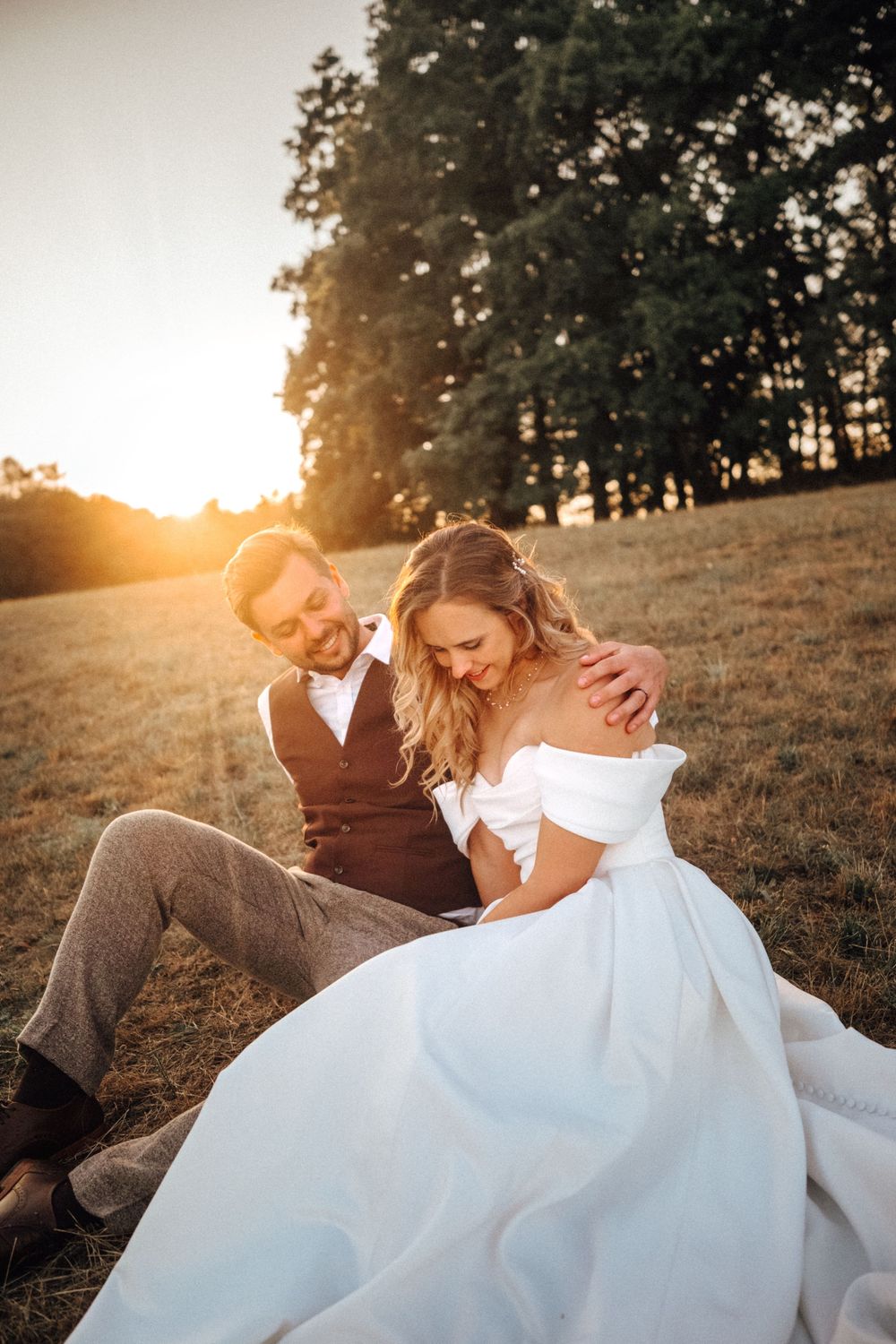 Bride and groom sitting together in golden sunset field, bride in white off-shoulder gown, groom in gray vest suit.