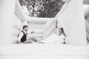 Bride and groom sitting together in a white inflatable bounce house, laughing in a fun black and white wedding photo.