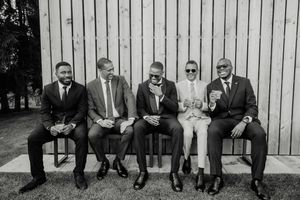 Five men in formal suits sitting on a bench outdoors, laughing and socializing in a black and white photo.